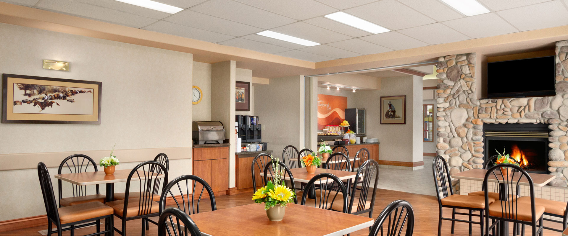 Large panoramic view of the brightly lit breakfast room at Days Inn, Red Deer featuring clean eating tables with comfortable chairs and stone fireplace.
